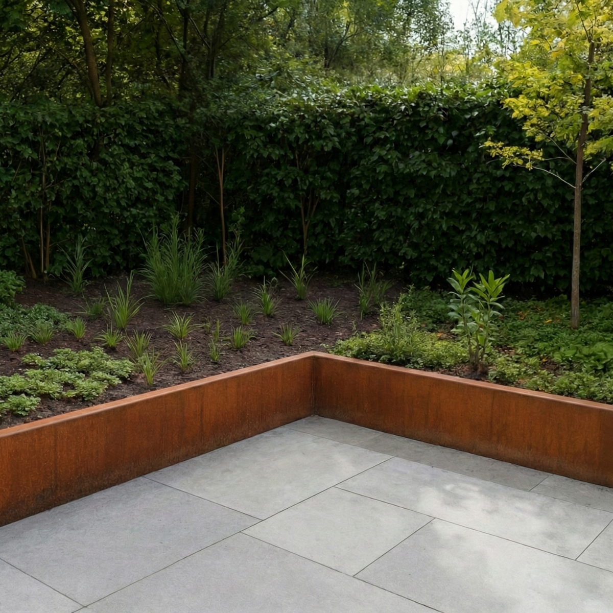 Lage border van cortenstaal langs een modern betegeld terras met diverse vaste planten en een groene haag.