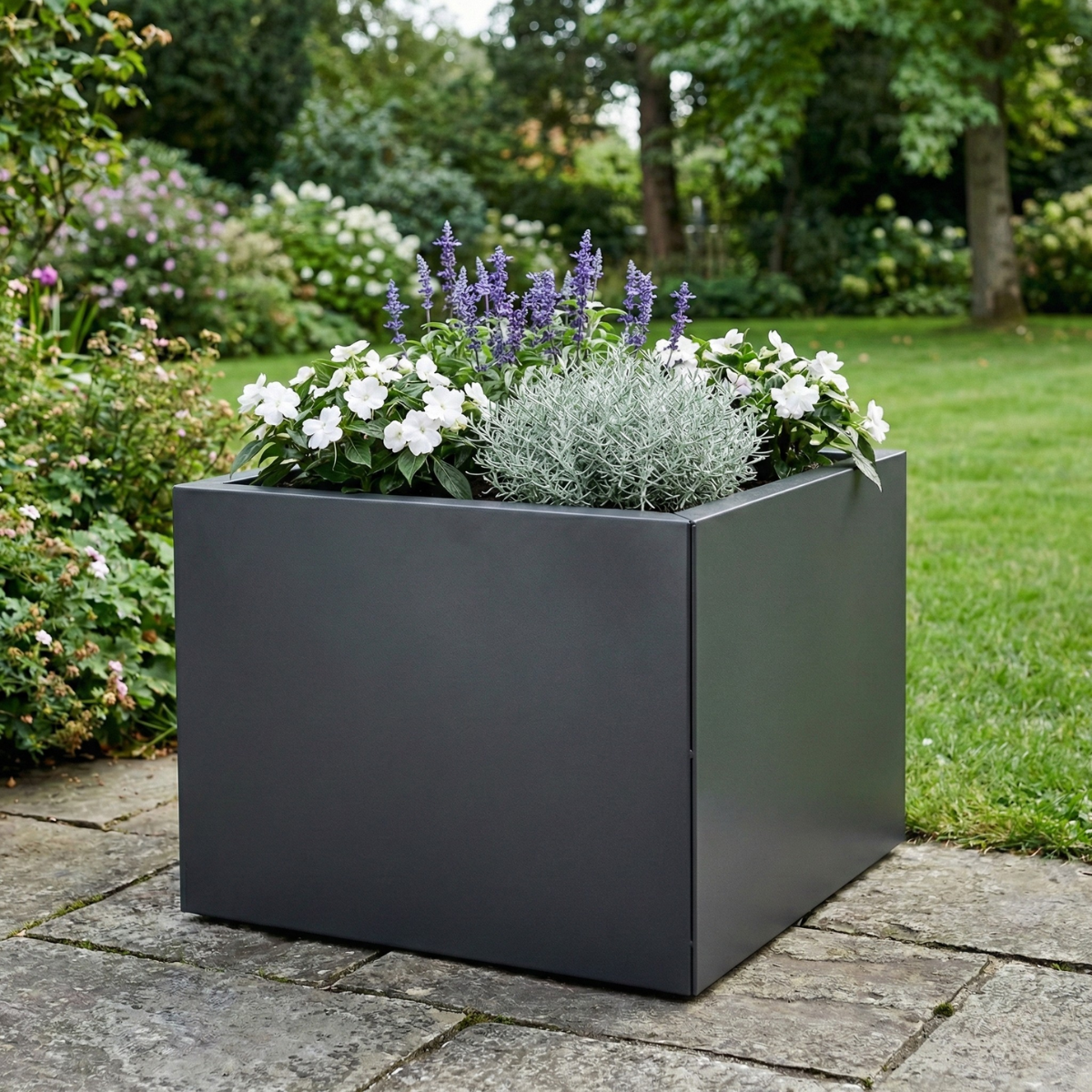 Grijze plantenbak, rijkelijk gevuld met sfeervolle paarse en witte bloemen en een zilverkleurig struikje, staand op een plavuizen pad in een groene tuin.