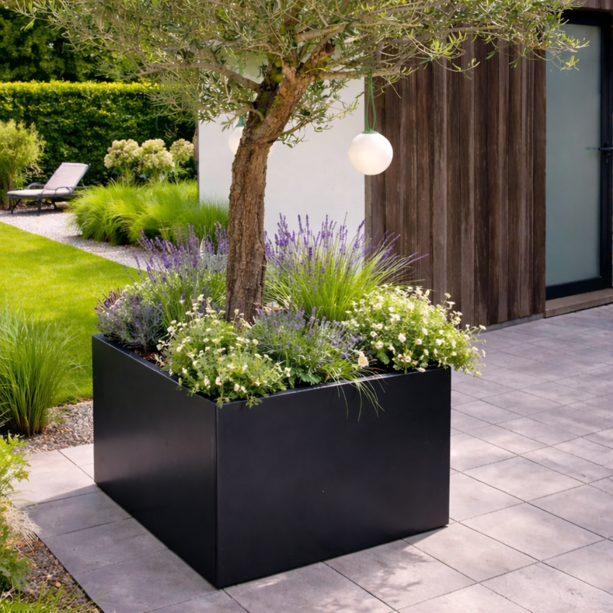 Grote zwarte vierkante plantenbak gevuld met een olijfboom paarse lavendel en witte bloemen op een betegeld terras in een sfeervolle moderne tuin.