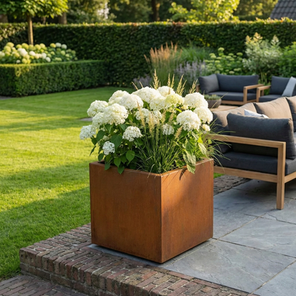 Vierkante Cortenstalen plantenbak met witte hortensia's en siergrassen op een modern terras bij een loungebank.