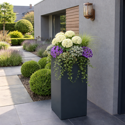 Hoge antracietkleurige plantenbak gevuld met witte hortensia's paarse bloemen en hangende klimop op een modern terras naast een strakke grijze muur.