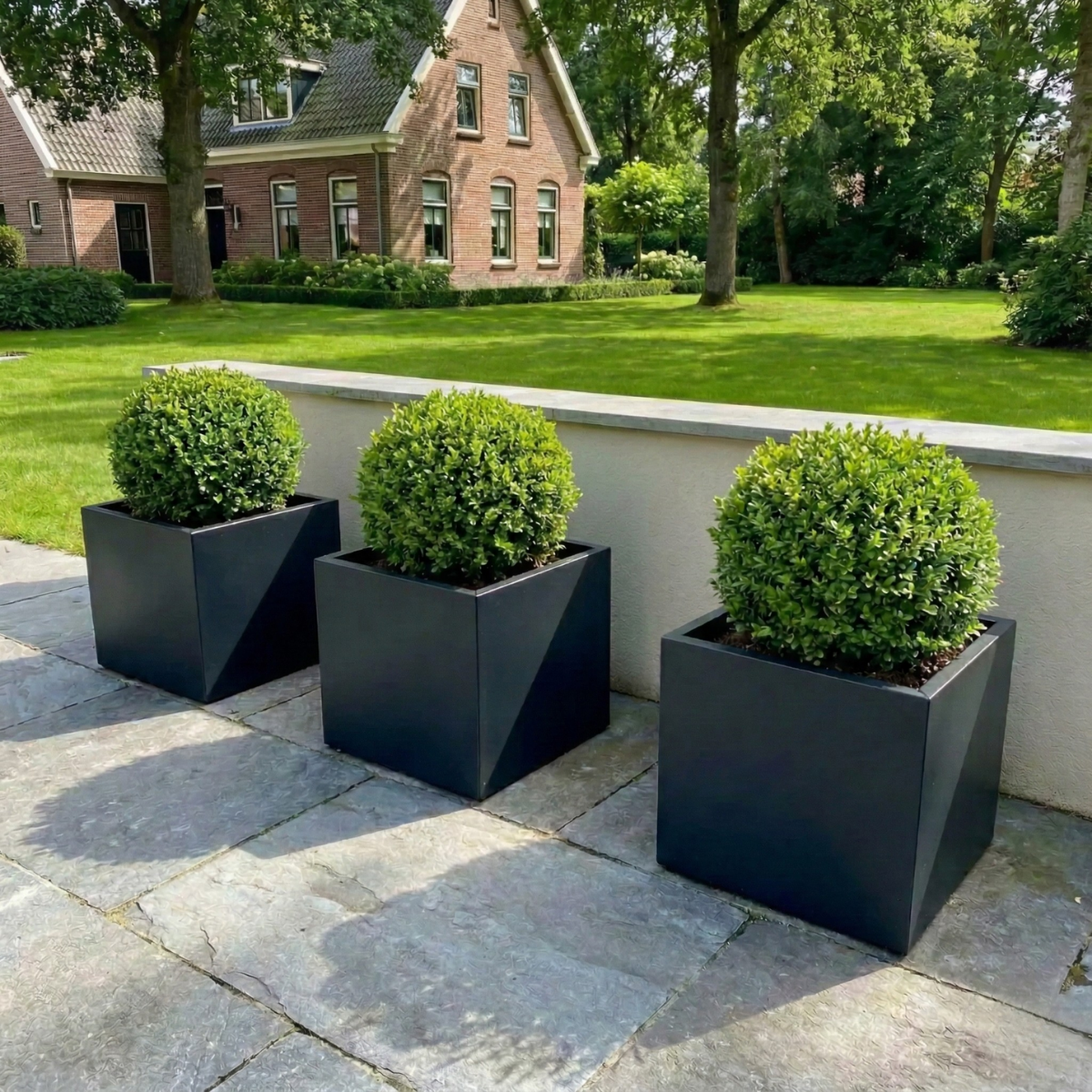 Hoge antracietgrijze vierkante plantenbak gevuld met een groen boompje op een betegeld terras naast een glazen pui van een modern wit huis.