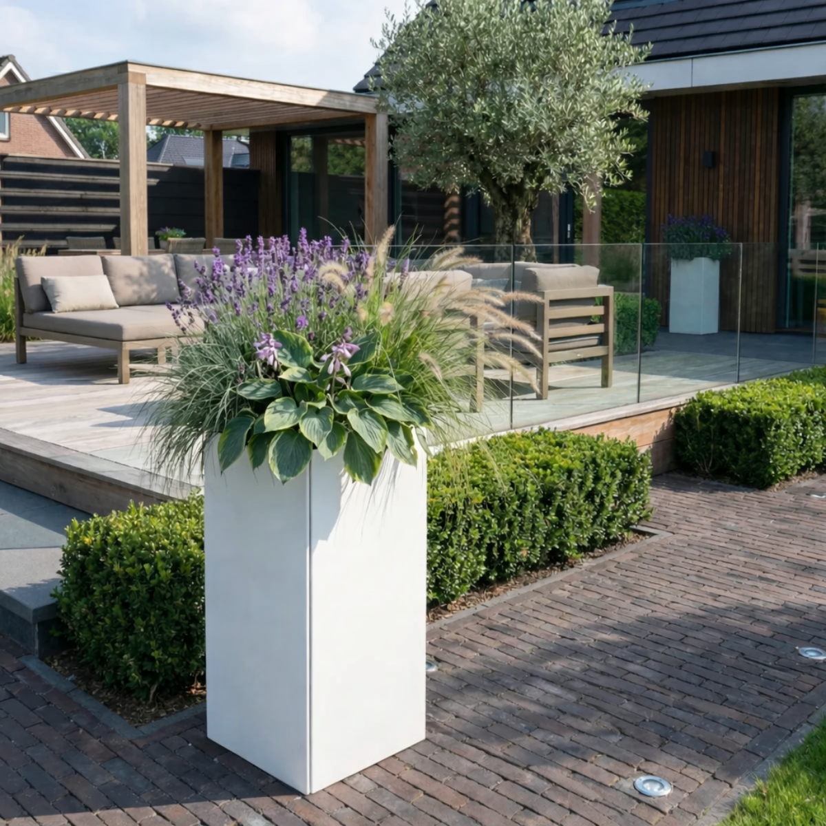 Hoge strakke witte plantenbak gevuld met paarse lavendel siergrassen en hosta op een klinkerpad in een sfeervolle moderne tuin met loungeset en pergola.