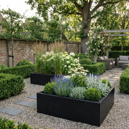 Twee grote zwarte rechthoekige plantenbakken gevuld met paarse bloemen witte zonnehoed en siergrassen in een sfeervolle tuin met grind en een oude bakstenen muur.