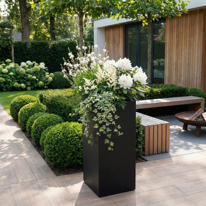 Hoge zwarte rechthoekige plantenbak met weelderige witte beplanting en hangende klimop op een modern terras met lichte tegels.
